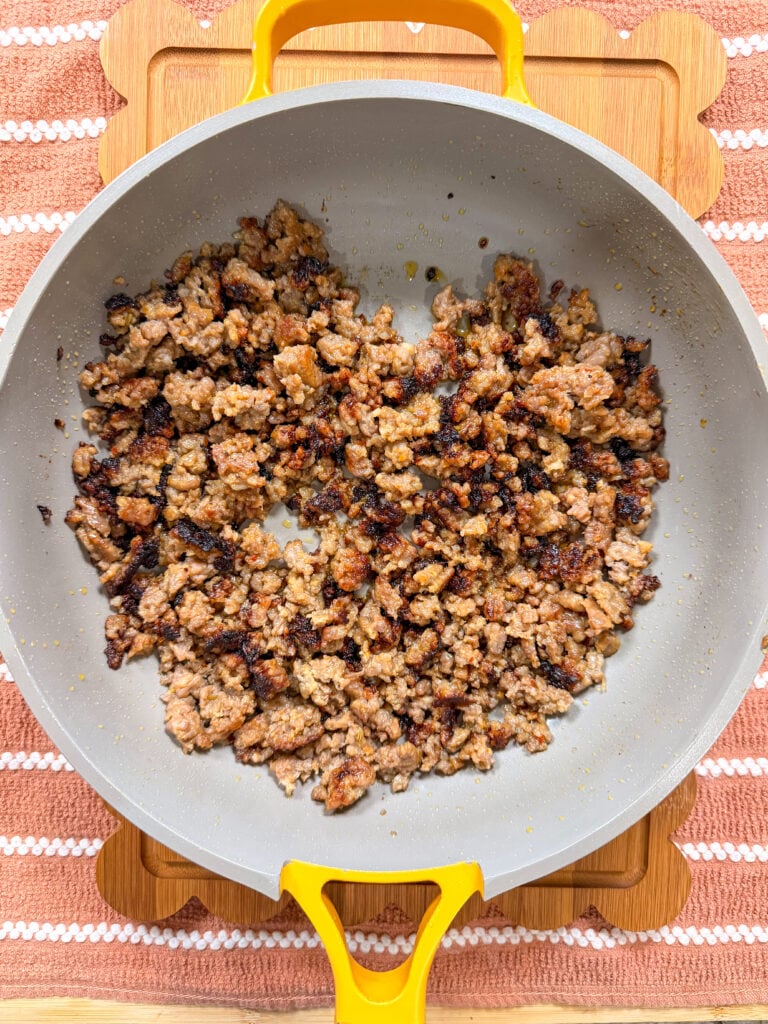 Cooked ground sausage in a yellow-handled skillet sits on a wooden board with a pink and white striped cloth underneath. The sausage is browned and slightly crispy.
