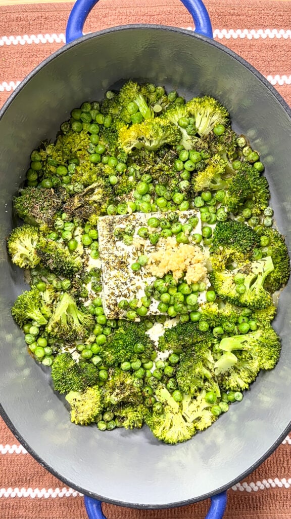 A blue oval baking dish with broccoli florets, green peas, a block of feta cheese, and minced garlic, all seasoned and ready to roast. The dish is placed on a rust-colored cloth with white stripes.