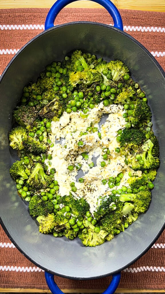 Roasted broccoli and peas with crumbled cheese and herbs in a large oval blue cast iron dish, placed on a patterned brown and white cloth.