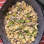 A close-up of orecchiette pasta with ground meat, peas, cheese, and a green herb sauce in a dark oval dish on a striped surface.