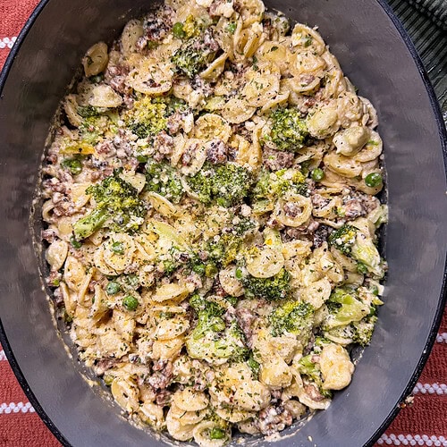 A close-up of orecchiette pasta with ground meat, peas, cheese, and a green herb sauce in a dark oval dish on a striped surface.