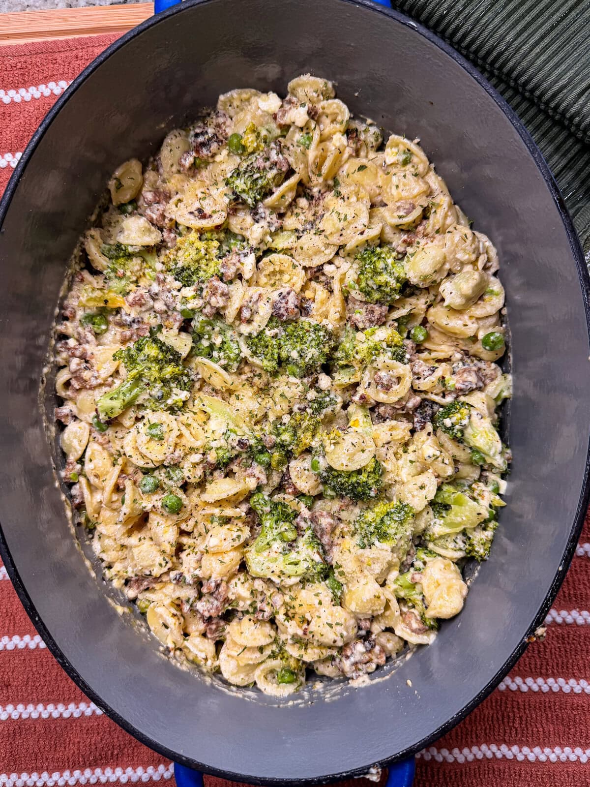 A close-up of orecchiette pasta with ground meat, peas, cheese, and a green herb sauce in a dark oval dish on a striped surface.
