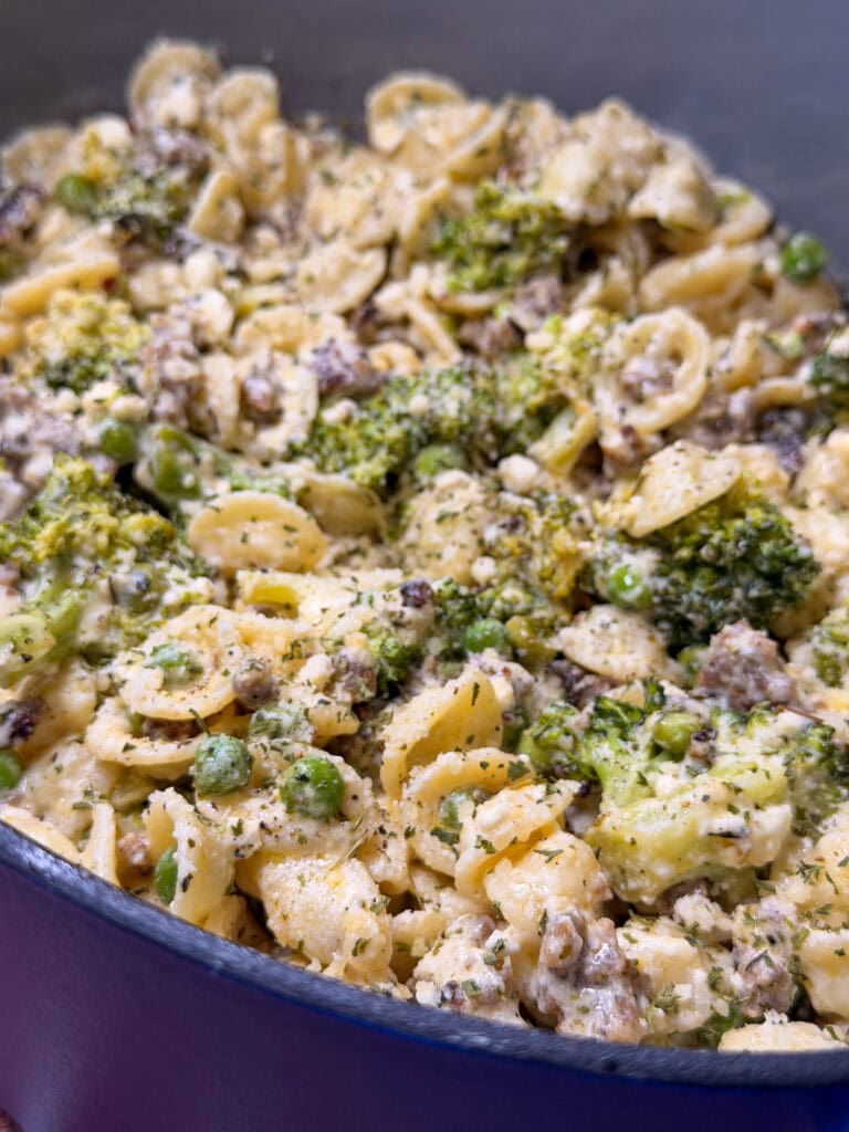 A close-up of a creamy pasta dish with orecchiette, ground meat, broccoli, green peas, and herbs in a large blue pot.
