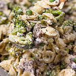 A wooden spoon scoops creamy pasta with orecchiette, broccoli, peas, ground meat, and ricotta cheese, all mixed together.