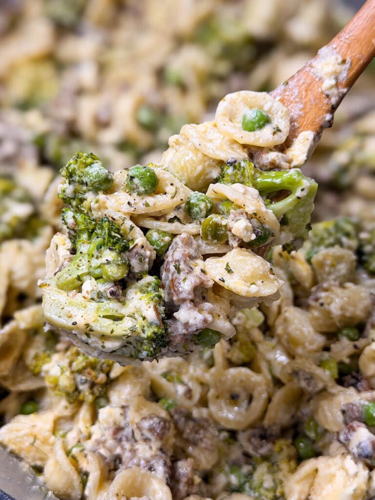 A wooden spoon scoops creamy pasta with orecchiette, broccoli, peas, ground meat, and ricotta cheese, all mixed together.