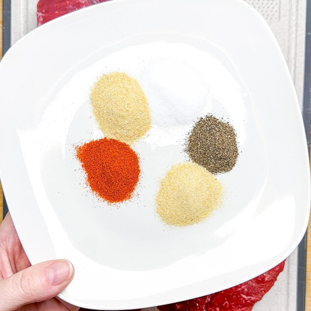 A hand holds a white plate with five piles of spices: white salt, black pepper, red paprika, yellow onion powder, and off-white garlic powder. A piece of raw meat is partially visible underneath.