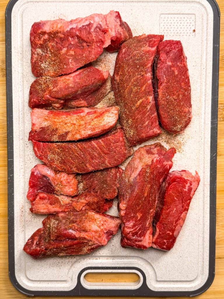 Raw beef pieces seasoned with spices are arranged on a rectangular cutting board with a handle, sitting on a wooden surface.