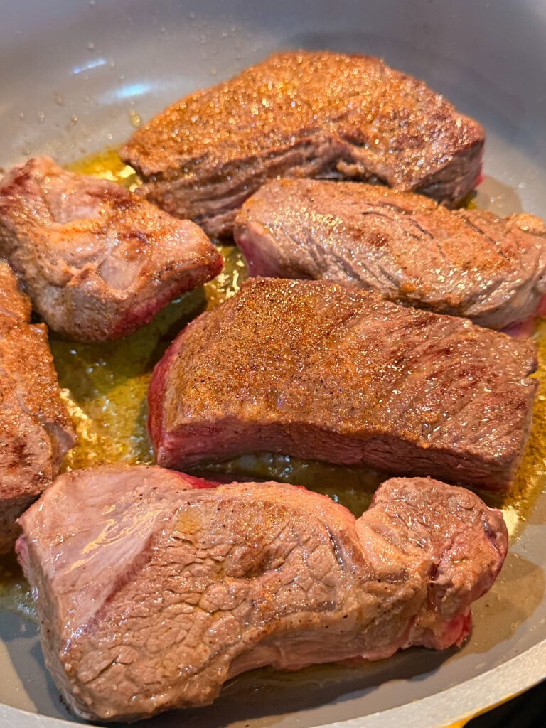 Chunks of beef short ribs searing in a pan, with a golden-brown crust forming and juices sizzling around the meat.