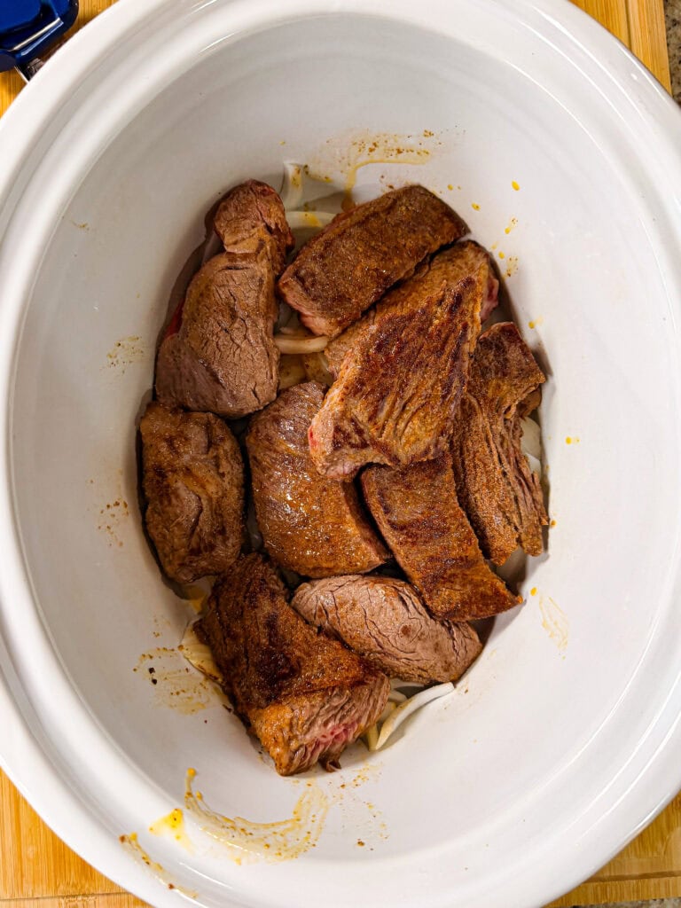 Chunks of browned, seasoned beef are arranged in a white slow cooker, sitting on top of a bed of sliced onions. The slow cooker is placed on a wooden surface.