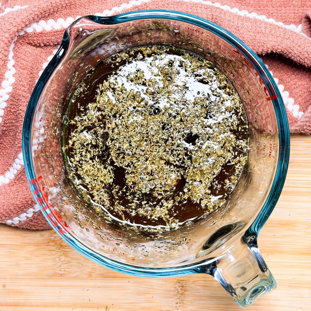 A glass measuring cup filled with a brown liquid and dried herbs sits on a wooden surface next to a textured, pink and white towel.