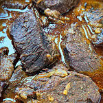Close-up of several pieces of braised beef in a rich, brown sauce. The meat is tender and coated with seasonings, with visible juices and oil in the dish.
