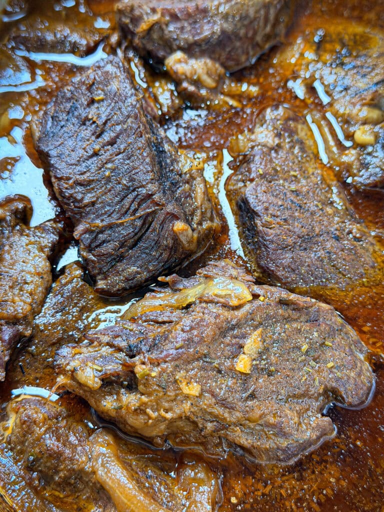 Close-up of several pieces of braised beef in a rich, brown sauce. The meat is tender and coated with seasonings, with visible juices and oil in the dish.