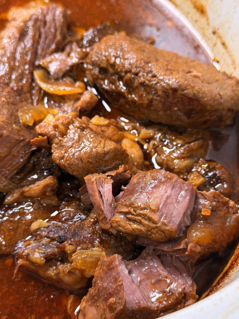 Close-up of tender, shredded pot roast beef in a rich brown sauce with visible onions in a white dish.