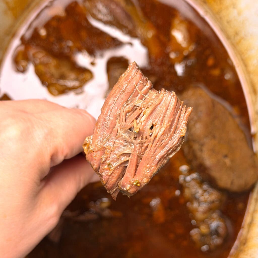 A hand holds up a piece of tender, shredded roast beef above a pot filled with more beef and rich brown cooking juices.