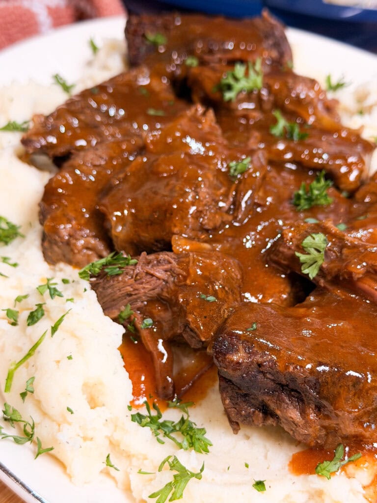 Braised beef with brown gravy served over mashed potatoes, garnished with chopped parsley on a white plate.