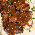 Braised beef pot roast with brown gravy served over mashed potatoes, garnished with chopped parsley on a white plate.