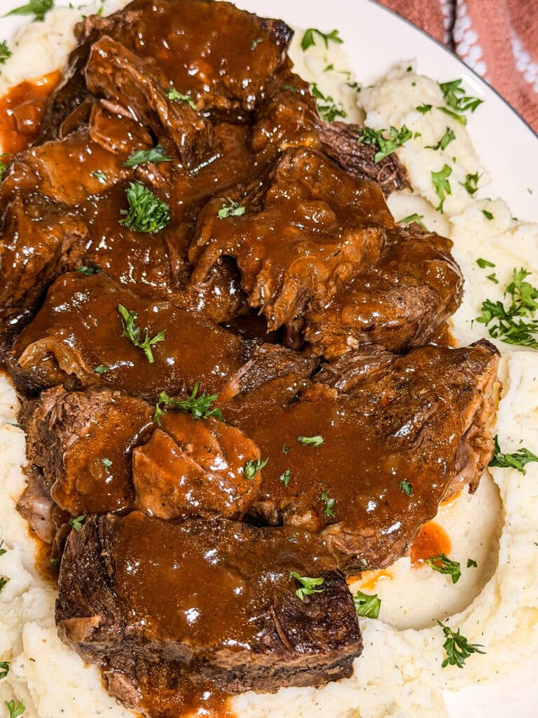Braised beef pot roast with brown gravy served over mashed potatoes, garnished with chopped parsley on a white plate.