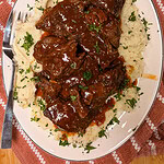 Plate of braised beef in brown sauce served over mashed potatoes, garnished with chopped parsley. A fork rests on the side of the plate, which is set on a red patterned cloth.