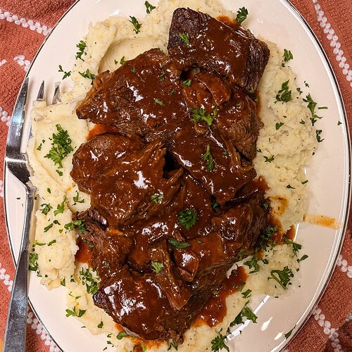 Plate of braised beef in brown sauce served over mashed potatoes, garnished with chopped parsley. A fork rests on the side of the plate, which is set on a red patterned cloth.