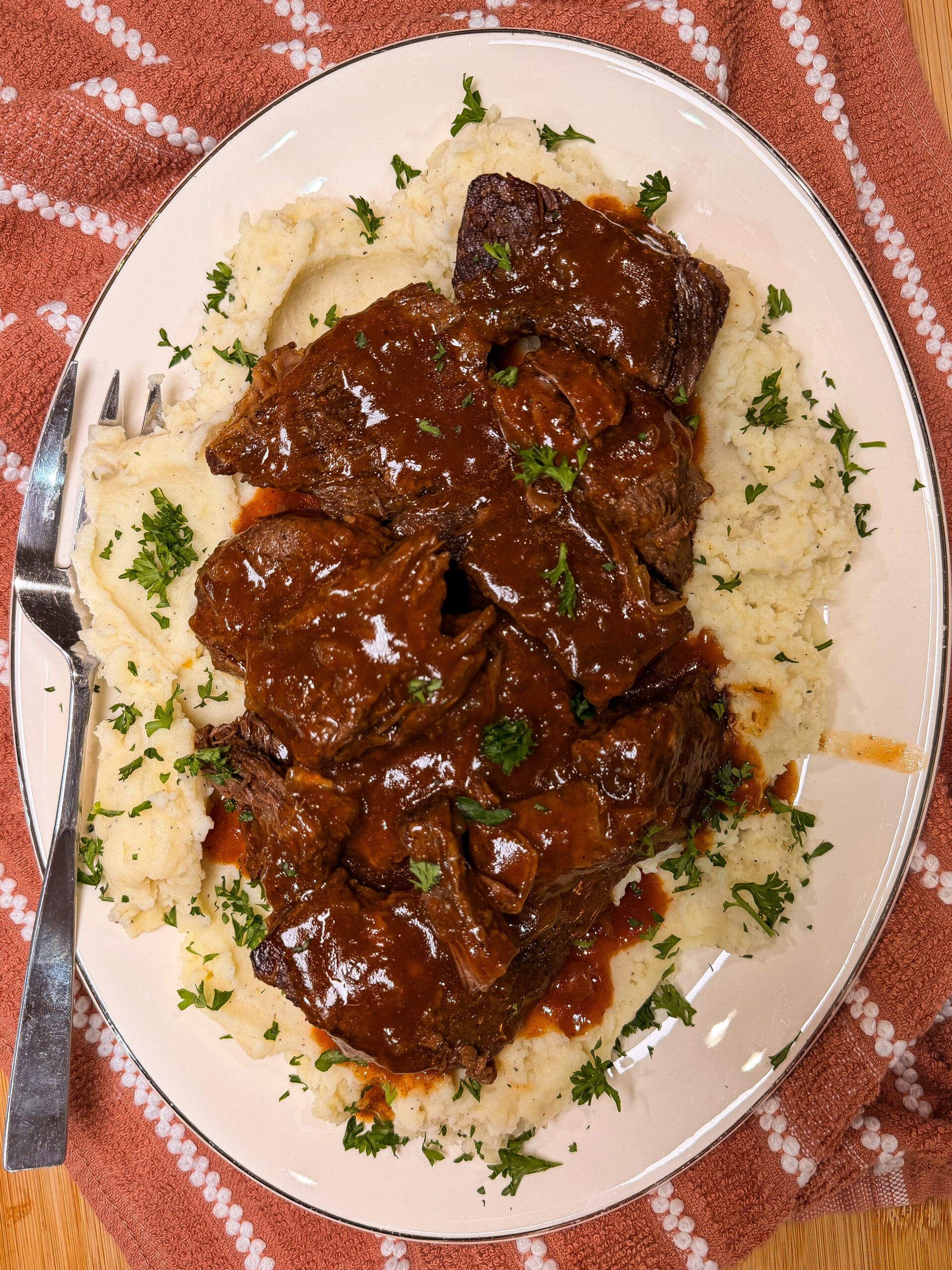 Plate of braised beef in brown sauce served over mashed potatoes, garnished with chopped parsley. A fork rests on the side of the plate, which is set on a red patterned cloth.