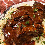 Pot roast covered in rich brown gravy sits atop a bed of mashed potatoes, garnished with chopped parsley, on a white plate with a steak knife nearby.