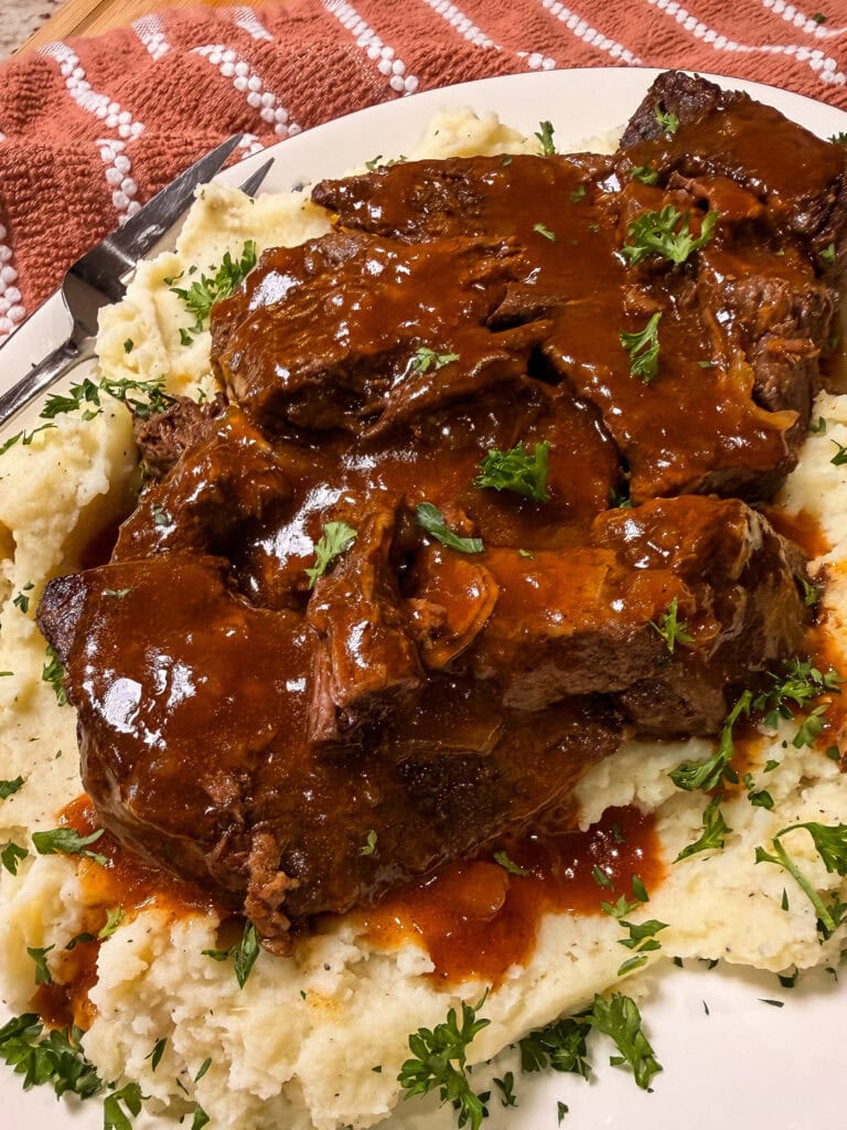Pot roast covered in rich brown gravy sits atop a bed of mashed potatoes, garnished with chopped parsley, on a white plate with a steak knife nearby.