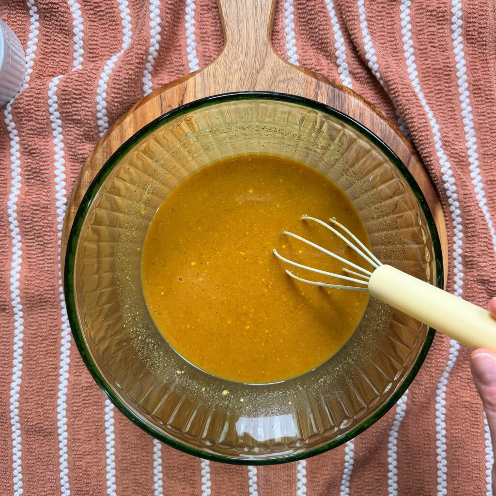 A hand holds a whisk in a glass bowl filled with golden-orange batter, set on a wooden board over a pink striped towel.