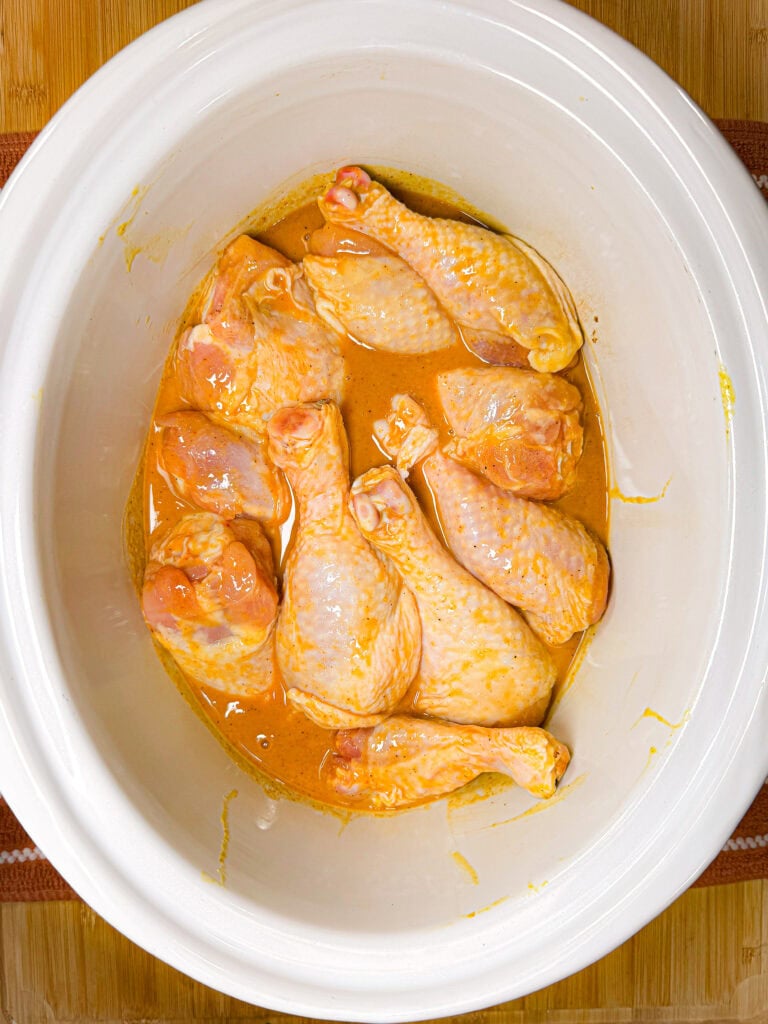 Raw chicken drumsticks coated in a yellow marinade sit arranged in a white oval slow cooker, ready to be cooked. The surface beneath is light brown wood.