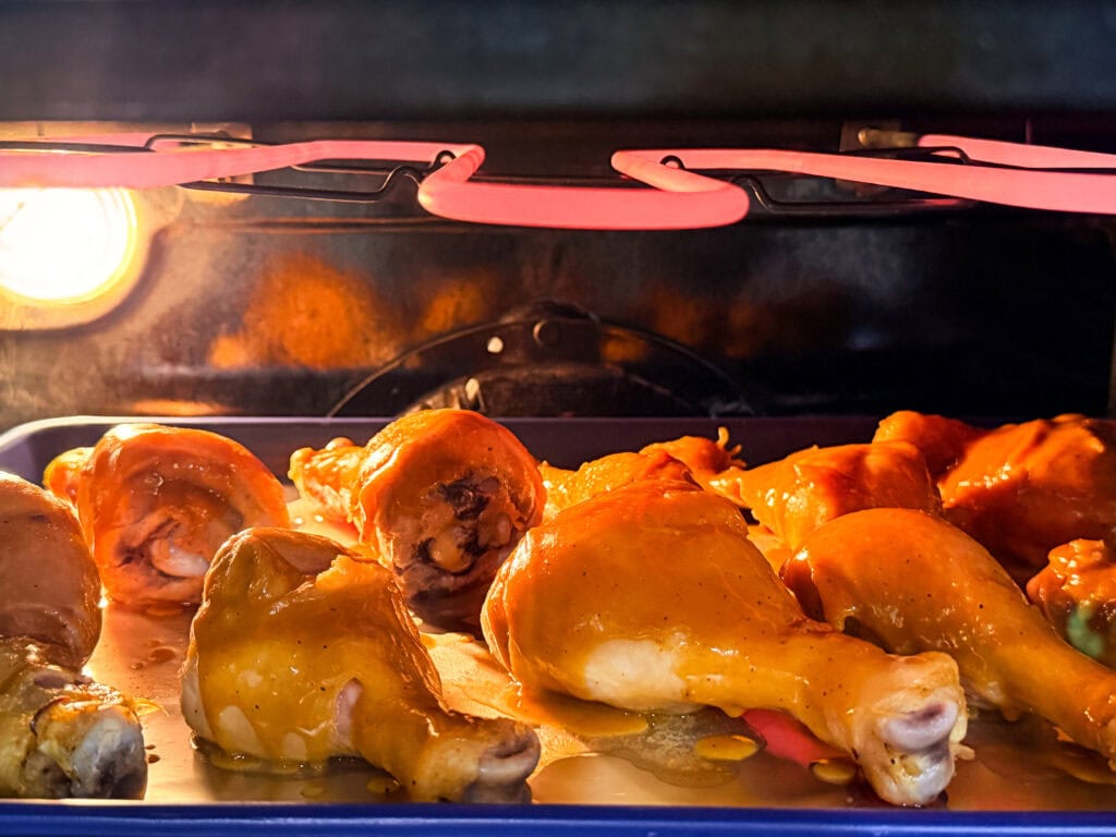 Chicken drumsticks coated in sauce are baking on a tray in an oven, with a warm yellow oven light illuminating the scene. The oven heating element above is glowing red-hot.