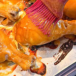 A close-up of a pink silicone brush spreading sauce on a baked chicken drumstick, surrounded by other saucy drumsticks on a baking sheet.