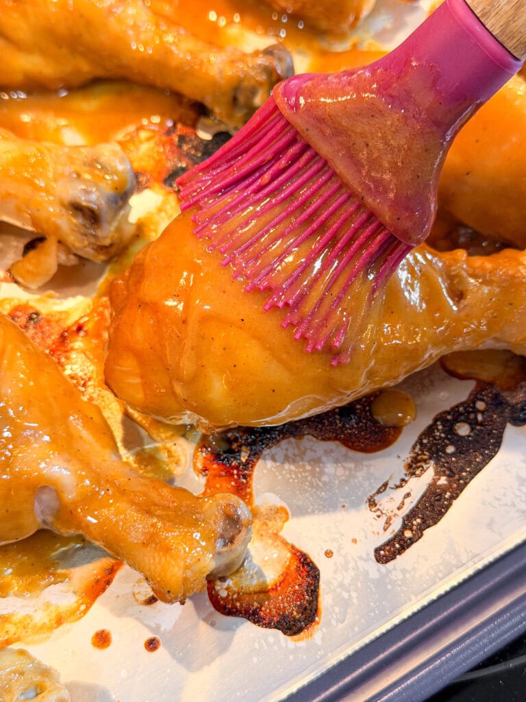 A close-up of a pink silicone brush spreading sauce on a baked chicken drumstick, surrounded by other saucy drumsticks on a baking sheet.