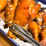 A pair of metal tongs holds a glazed, baked chicken drumstick in the foreground, with several more drumsticks on a tray in the background.