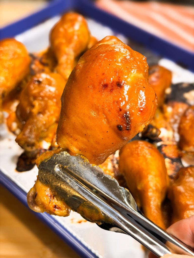 A pair of metal tongs holds a glazed, baked chicken drumstick in the foreground, with several more drumsticks on a tray in the background.