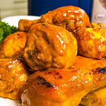 Close-up of several pieces of baked or grilled chicken coated in a golden, glossy sauce, served on a white plate with a blue rim. Part of a leafy green garnish is visible in the background.