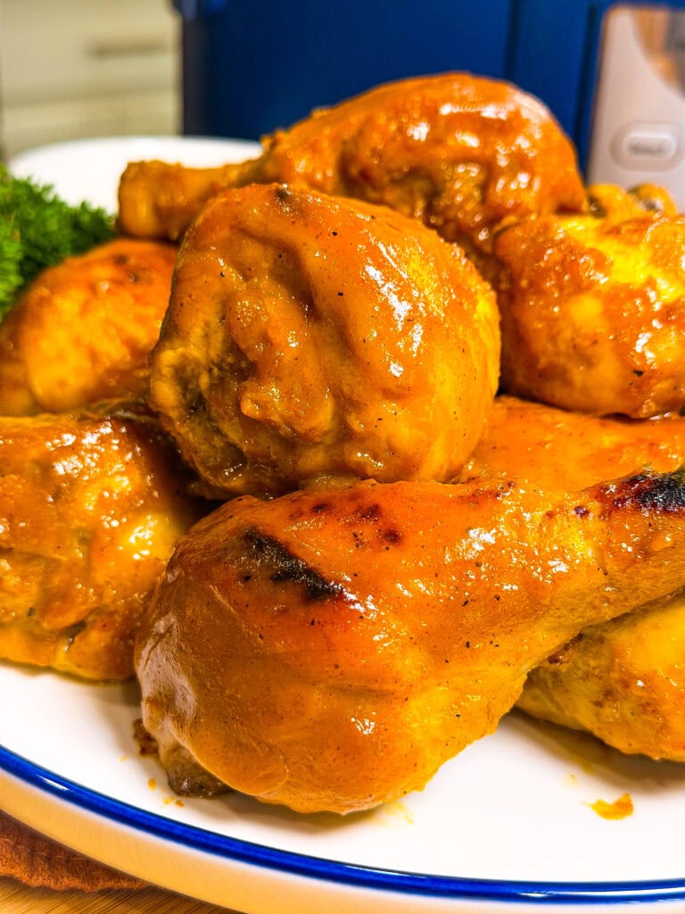 Close-up of several pieces of baked or grilled chicken coated in a golden, glossy sauce, served on a white plate with a blue rim. Part of a leafy green garnish is visible in the background.