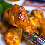 A close-up of metal tongs holding a saucy, grilled chicken drumstick over a plate of more chicken, with a bowl of dipping sauce and greens blurred in the background.