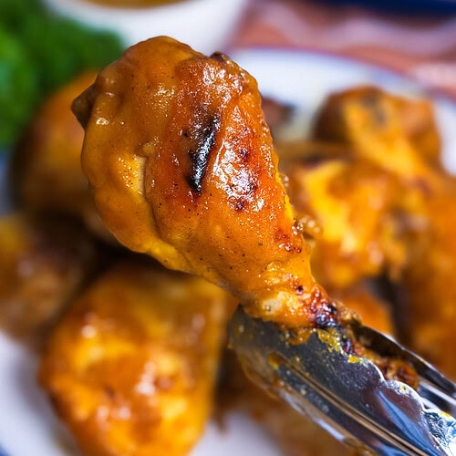 A close-up of metal tongs holding a saucy, grilled chicken drumstick over a plate of more chicken, with a bowl of dipping sauce and greens blurred in the background.
