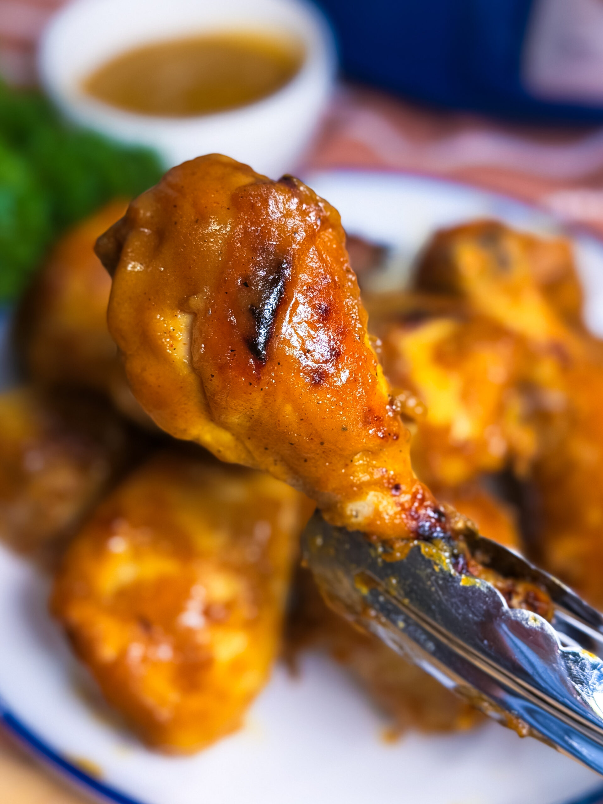 A close-up of metal tongs holding a saucy, grilled chicken drumstick over a plate of more chicken, with a bowl of dipping sauce and greens blurred in the background.