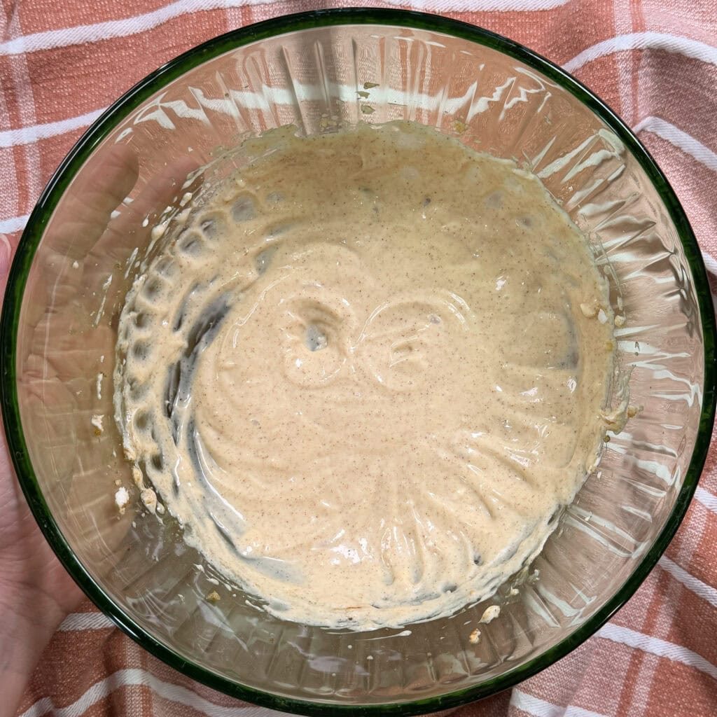 A glass bowl with a thick, beige batter, partially mixed, sits on a pink-and-white striped cloth. The bowl shows swirls in the batter and some remains are stuck around the sides.