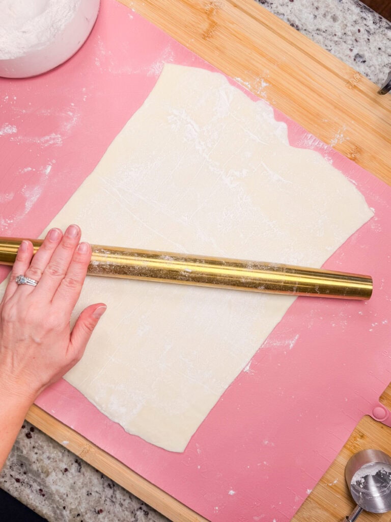 A hand uses a gold rolling pin to flatten dough on a pink surface. Flour and baking tools are nearby on a wooden board and granite countertop.