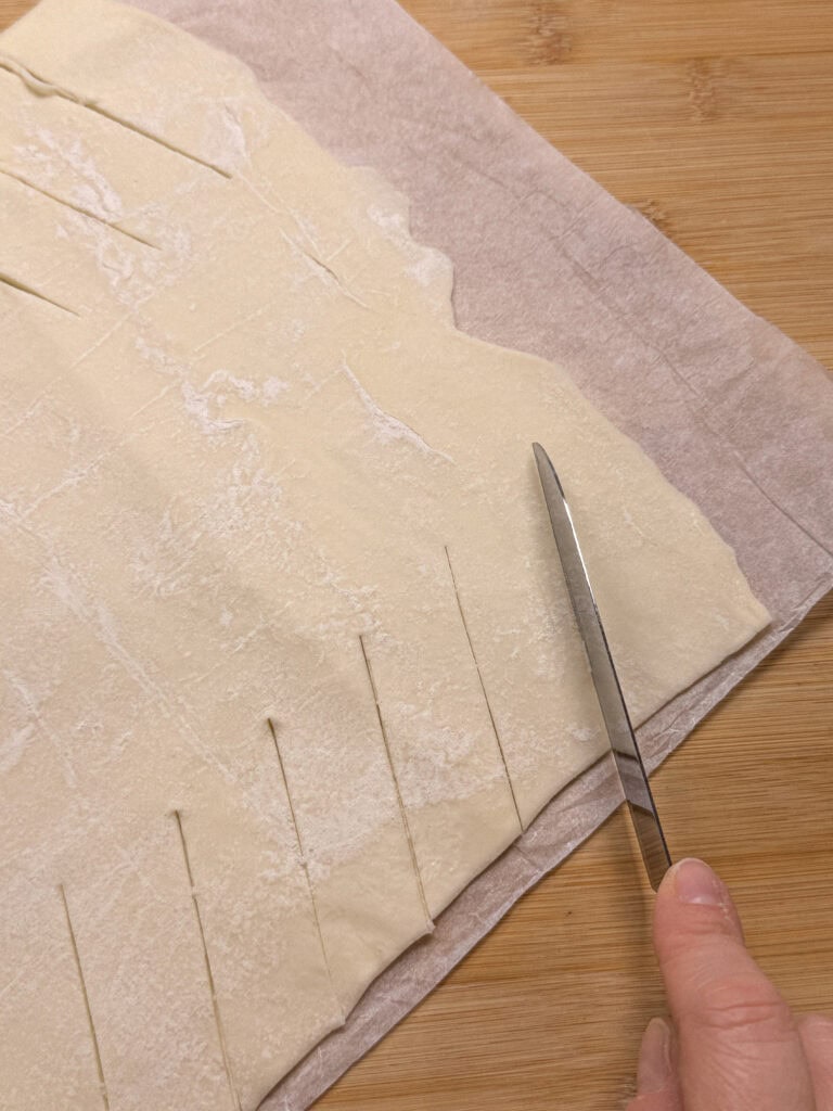 A hand uses a knife to cut parallel slits into a sheet of puff pastry dough placed on parchment paper, on a wooden surface.