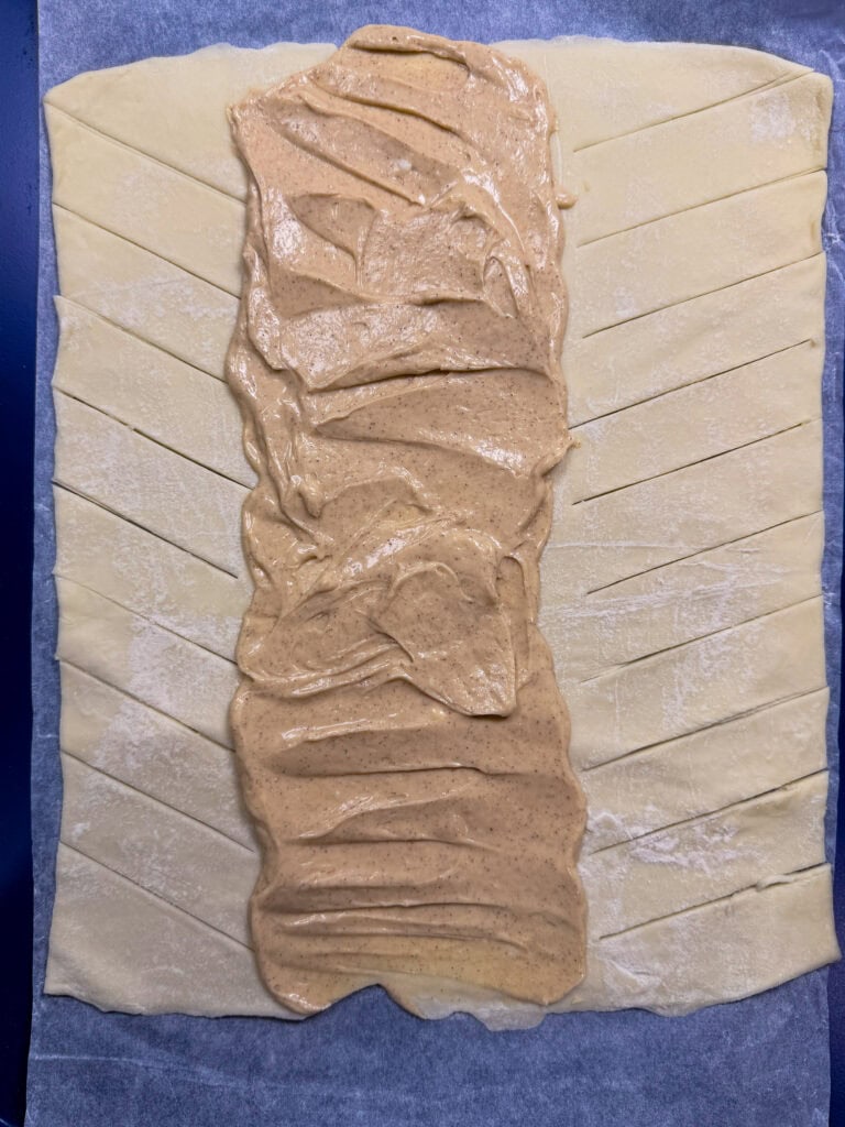 A sheet of pastry dough with diagonal slits on both sides, filled in the center with a creamy nut butter mixture, ready to be braided or folded before baking.