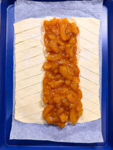 Unbaked braided pastry on parchment, with peach filling in the center and diagonal slits cut into the dough on each side, ready to be folded over the filling.