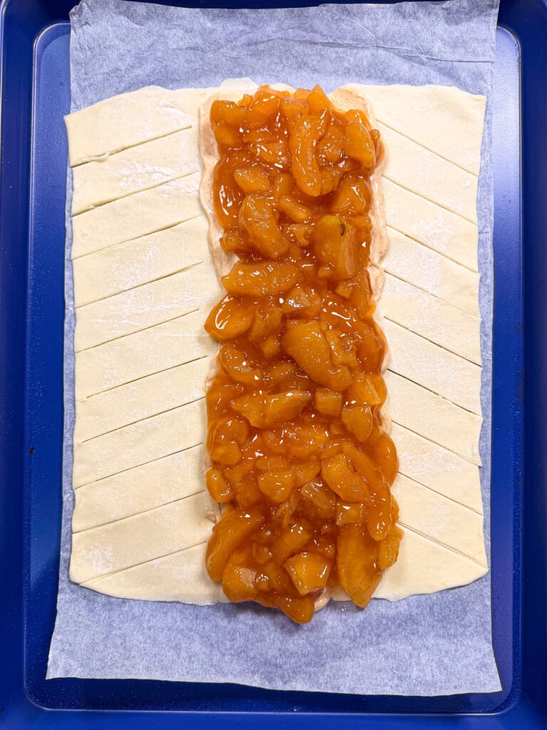 Unbaked braided pastry on parchment, with peach filling in the center and diagonal slits cut into the dough on each side, ready to be folded over the filling.