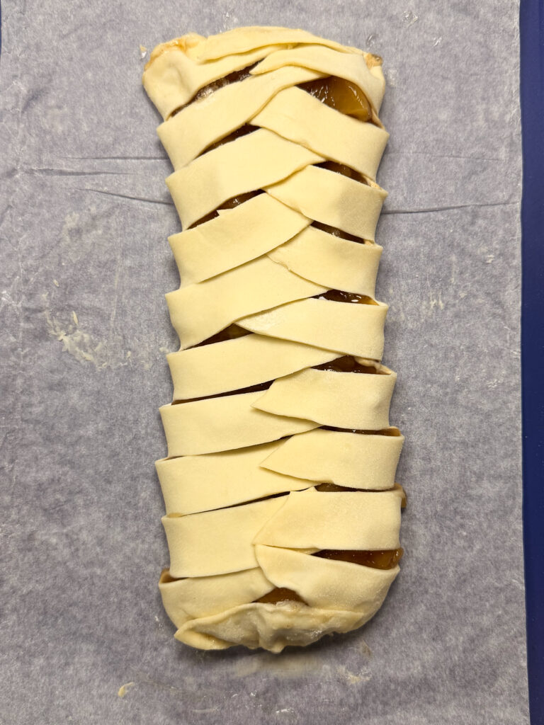 Unbaked pastry braid with a fruit filling, made from strips of dough woven over each other, resting on a sheet of parchment paper.