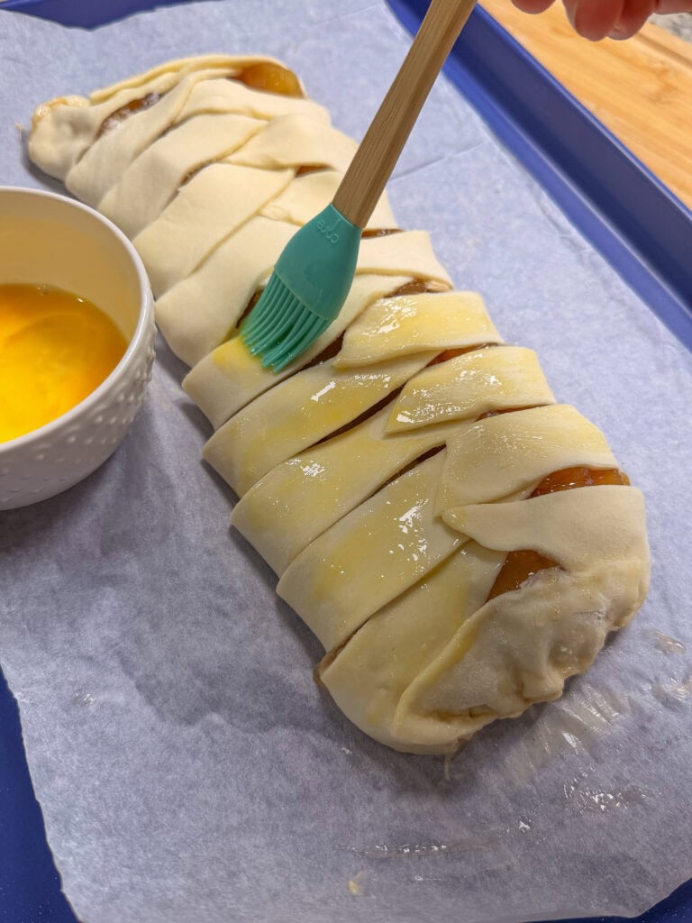 A hand uses a green brush to apply egg wash to a braided pastry on parchment paper, with a small bowl of egg wash nearby.