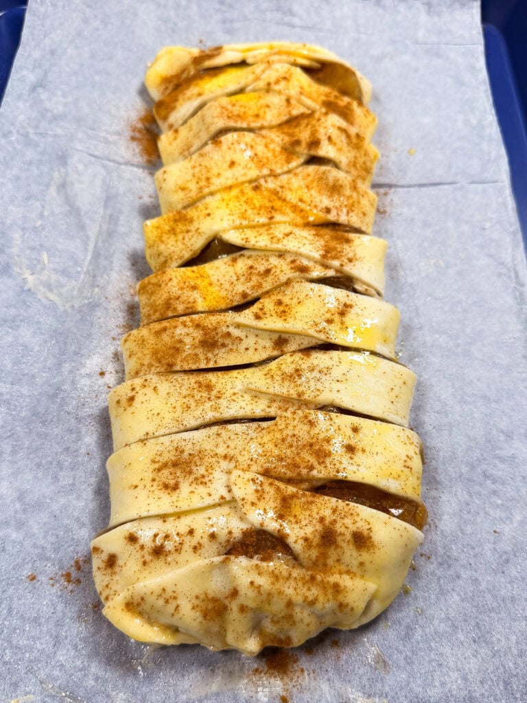 Unbaked braided pastry with slits on top, sprinkled with cinnamon, sits on parchment paper, ready for baking.