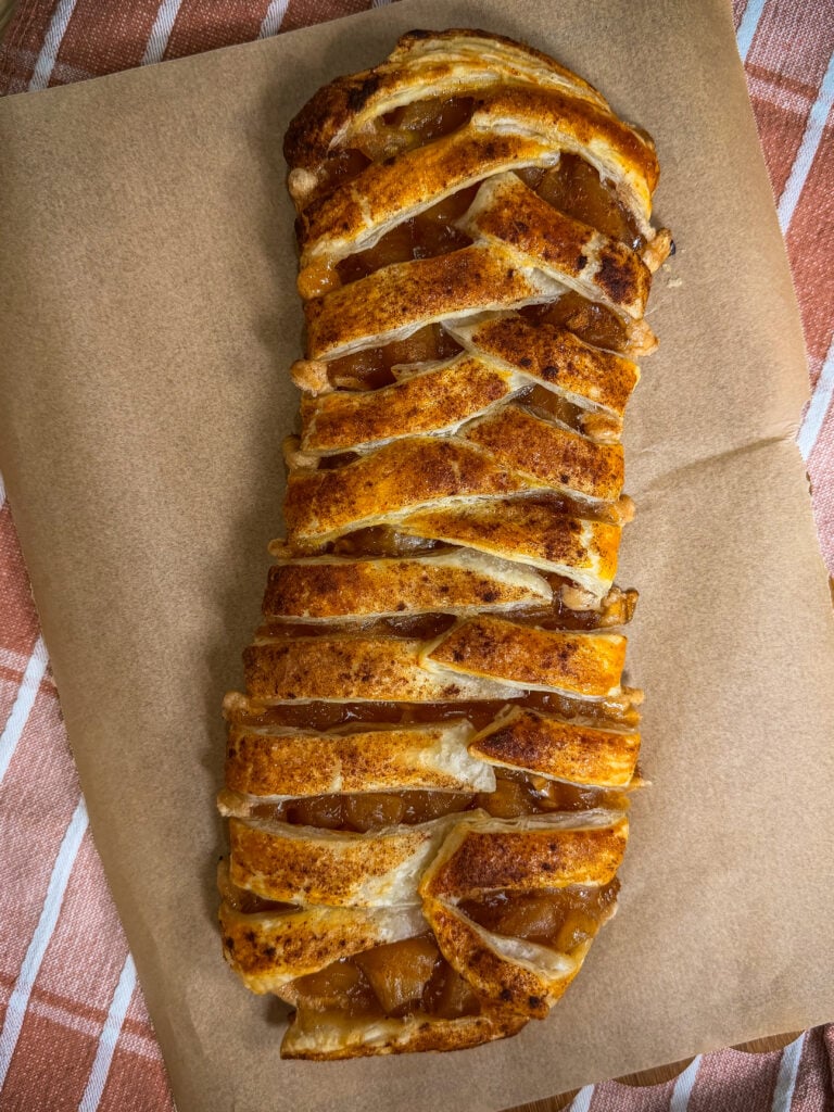 A golden-brown apple strudel with a lattice crust sits on parchment paper, revealing chunks of apple filling. The strudel rests on a striped cloth background.