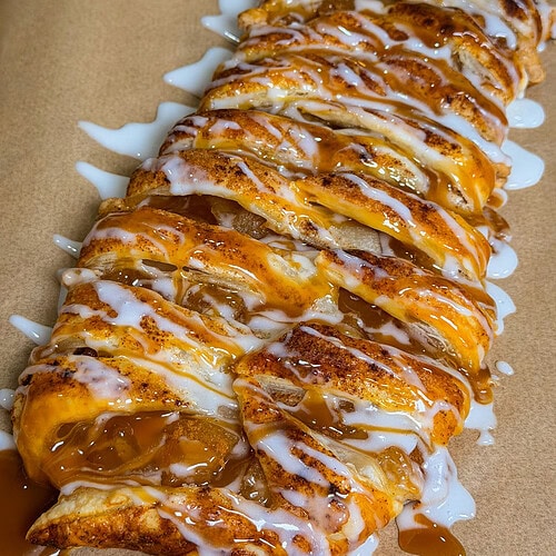 A braided pastry filled with cinnamon and apples, drizzled with white icing and caramel sauce, sits on brown parchment paper.