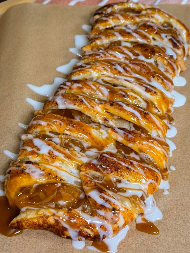 A braided pastry filled with cinnamon and apples, drizzled with white icing and caramel sauce, sits on brown parchment paper.